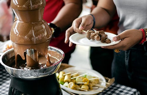 Hands dipping fruit into a chocolate fountain at a party.