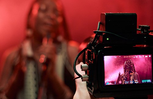 Camera filming a DJ at a party with lighting and crowd.